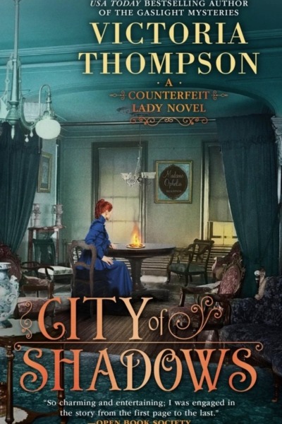 City of Shadows by Victoria Thompson book cover shows a well-dressed young woman with red hair in an early 1900s room, sitting at at table with a gas lamp burning on it. A sign on the wall says "Madame Ophelia - Readings"