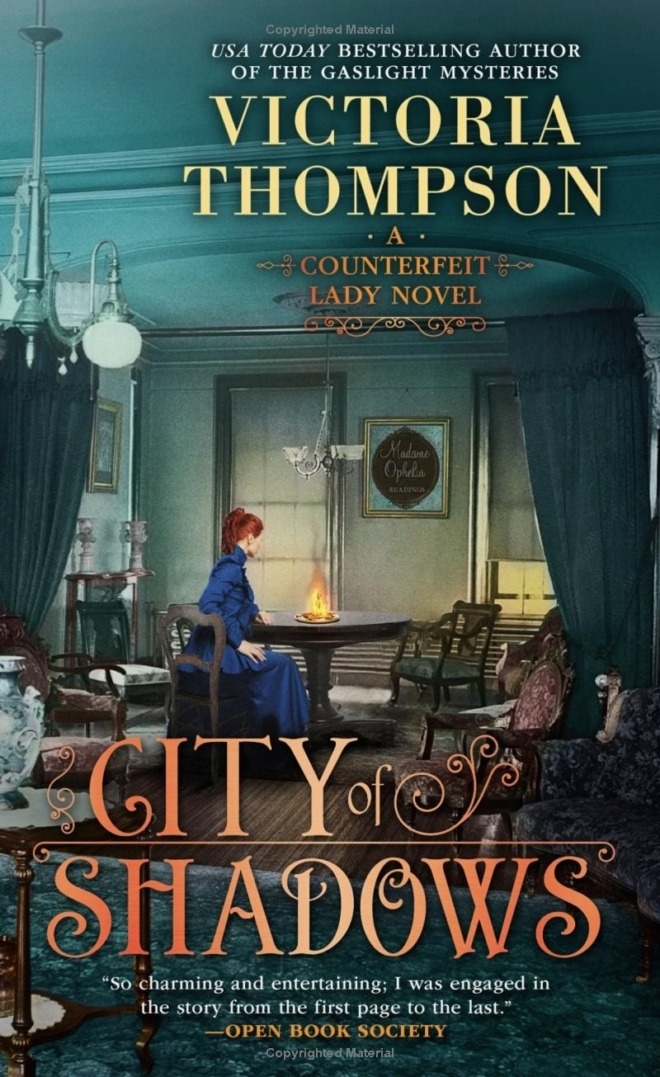 City of Shadows by Victoria Thompson book cover shows a well-dressed young woman with red hair in an early 1900s room, sitting at at table with a gas lamp burning on it. A sign on the wall says "Madame Ophelia - Readings"
