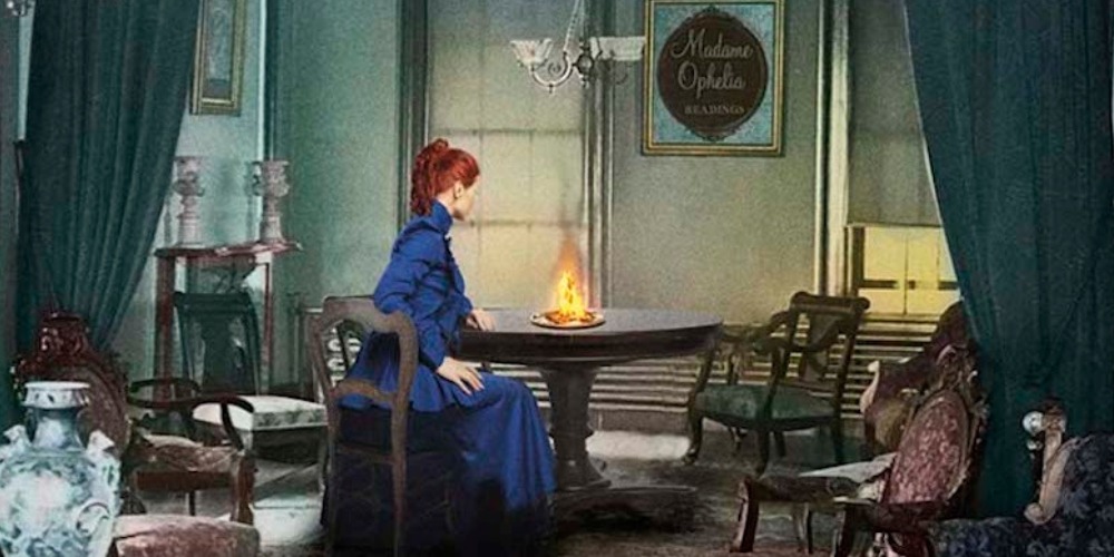 City of Shadows by Victoria Thompson book cover (detail) shows a well-dressed young woman with red hair in an early 1900s room, sitting at at table with a gas lamp burning on it. A sign on the wall says "Madame Ophelia - Readings"